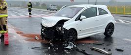 Widać biały uszkodzony samochód od przodu, jest na skrzyżowaniu