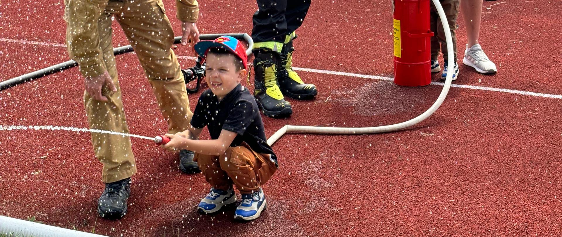 Dziecko leje wodę z hydronetki. Tata i brat pompują wodę ręcznie. Nad wszystkim czuwa strażak.