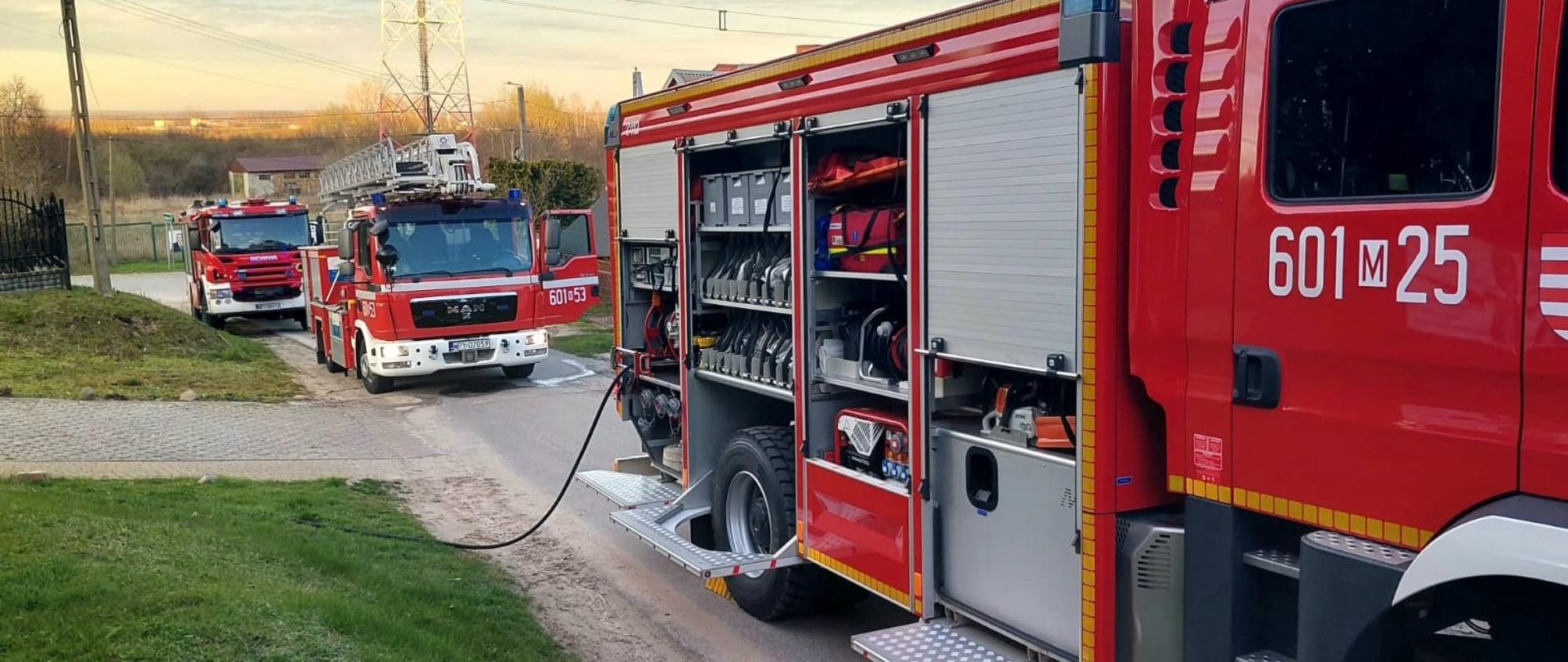 Zdjęcie przedstawia trzy czerwone wozy strażackie zaparkowane wzdłuż pobocza drogi lub ulicy. Na pierwszym planie, po prawej stronie, znajduje się duży wóz strażacki z otwartymi bocznymi żaluzjami, za którymi widać różnorodne wyposażenie, takie jak węże, drabiny i narzędzia. Na jego boku widoczny jest numer "601 25". Z tyłu tego pojazdu widać czarny wąż biegnący w dół. Za pierwszym wozem, w oddali, stoją dwa kolejne wozy strażackie, również czerwone. Jeden z nich ma na dachu drabinę, a na jego przedzie widnieje numer "601 53". Tło zdjęcia tworzy teren zabudowany z domami i drzewami oraz maszt radiowy lub telekomunikacyjny. Niebo jest jasne, częściowo zachmurzone, co sugeruje wczesny poranek lub późne popołudnie. Całość sceny wskazuje na gotowość operacyjną.