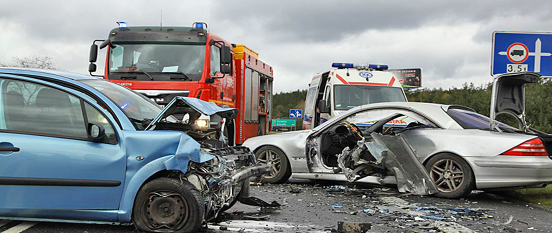 Fotografia przedstawia dwa uszkodzone samochody osobowe na skutek wypadku drogowego. W tle widać pojazd straży pożarnej oraz karetkę.