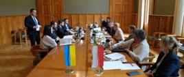 Zdjęcie z boku, po obu stronach podłużnego czworokątnego stołu siedzą ludzie, na stole dwie małe flagi - polska i ukraińska.
