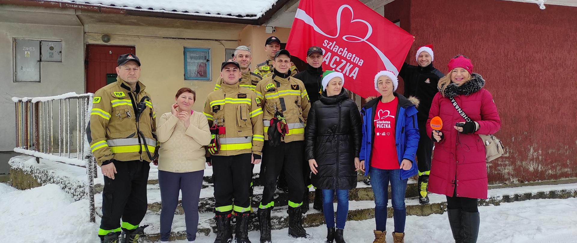 Na zdjęciu przy budynku pozują strażacy oraz wolontariuszki ze Szlachetnej Paczki. Strażacy trzymaja czerwona flagę z napisem Szlachetna Paczka.