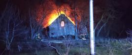 Na zdjęciu w porze nocnej widać dom w oddali, który jest w płomieniach. Przed pożarem widać strażaków oraz rozwinięta linię gaśniczą. Wokół miejsca zdarzenia rosną drzewa oraz krzaki.