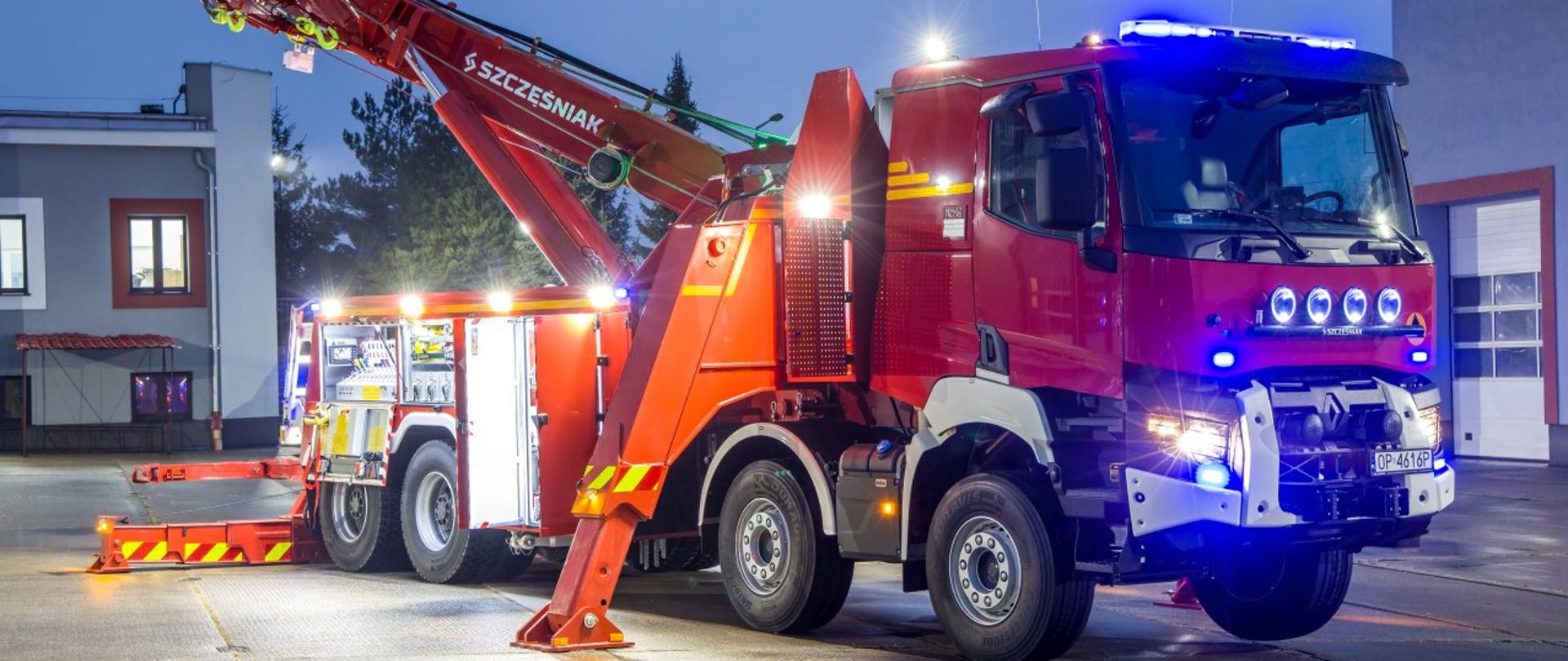 Przygotowany do pracy ciężki samochód ratownictwa drogowego typu rotator rozstawiony na placu KP PSP w Brzegu. W tle widoczny budynek Komendy Powiatowej PSP w Brzegu. Zdjęcie wykonane od przodu i prawej strony pojazdu w porze nocnej.