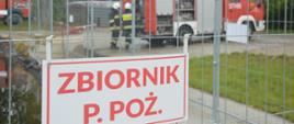 Akcja na składowisku odpadów. Na pierwszym planie zbiornik wody i opłotowanie, na którym zawieszona jest tabliczka "ZBIORNIK P.POŻ" W tle wóz straży pożarnej, przy którym stoi dwóch strażaków w białych hełmach oraz drzewa.