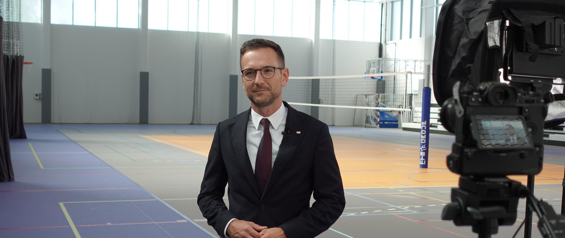 Minister Waldemar Buda stoi w sali sportowej liceum w Piastowie.