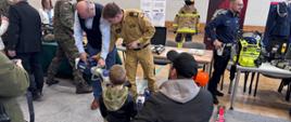 Na zdjęciu widać scenę z wydarzenia o charakterze edukacyjno-pokazowym, związanego ze służbami ratowniczymi i mundurowymi. W centrum znajduje się kilku dorosłych – część w umundurowaniu (wojskowym i strażackim), a także mężczyzna w stroju cywilnym – którzy prezentują sprzęt lub rozmawiają z dzieckiem stojącym przodem do nich. Obok dziecka klęczy opiekun, obserwując sytuację.
Po prawej stronie widoczny jest strażak w pełnym umundurowaniu oraz specjalistyczny sprzęt ratowniczy ustawiony na stole. W tle znajdują się elementy wyposażenia strażackiego, w tym manekin ubrany w strój ochronny oraz materiały informacyjne na roll-upach.
Całość przedstawia przyjazną, edukacyjną atmosferę – dorośli tłumaczą i pokazują dzieciom sprzęt, zachęcając do poznawania pracy służb ratunkowych.