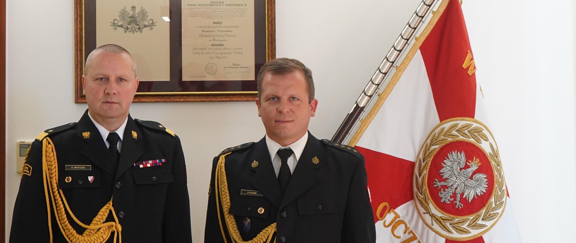 dwóch strażaków w mundurach ze sznurem stoi obok siebie, jeden z nich trzy,ma teczkę , w tle sztandar