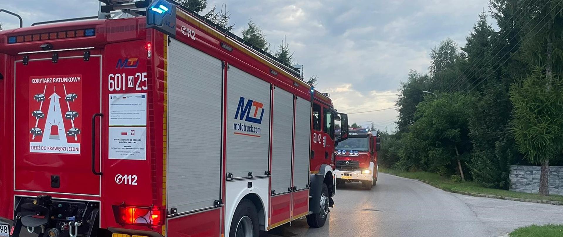 Zdjęcie przedstawia dwa czerwone wozy strażackie stojące na drodze. Na pierwszym planie widoczny jest tył większego wozu z numerem "601-25" oraz napisem "KORYTARZ RATUNKOWY" i instrukcją tworzenia korytarza życia. Na lewym boku pojazdu widnieje logo "MOTOTRUCK.com". W tle, za nim, widoczny jest przód drugiego wozu strażackiego. Droga jest mokra, a w oddali widać drzewa i budynki. Niebo jest pochmurne, co sugeruje wczesny ranek lub zmierzch.