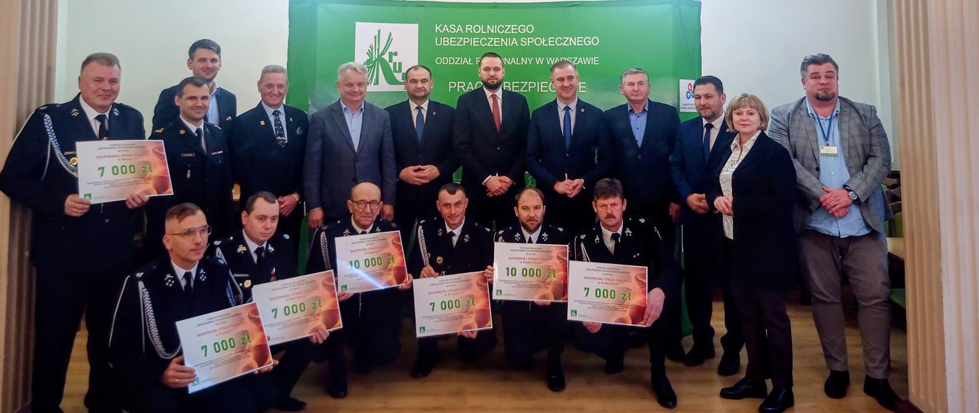 Grupa strażaków z wręczonymi promesami z kwotami przyznanego dofinasowania od 7000 do 10000 zł oraz przedstawiciele Starostwa Powiatowego w Grójcu Urzędów Gmin i Miast i Gmin oraz KRUS-u