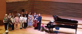 Grupa 11 dzieci i młodzieży stoi na estradzie w dwóch rzędach, po lewej stronie stoi kobieta, po prawej stronie stoi kobieta z wiolonczelą i mężczyzna. Po prawej stronie od Nich stoi fortepian, na krześle leżą kwiaty.