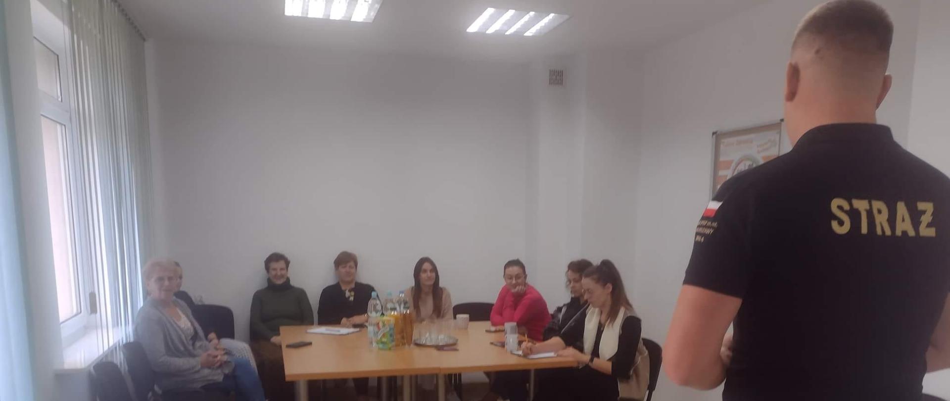 Pracownik straży pożarnej stoi przed kobietami siedzącymi przy stoliku. Na podłodze między nimi leżą dwa manekiny. 