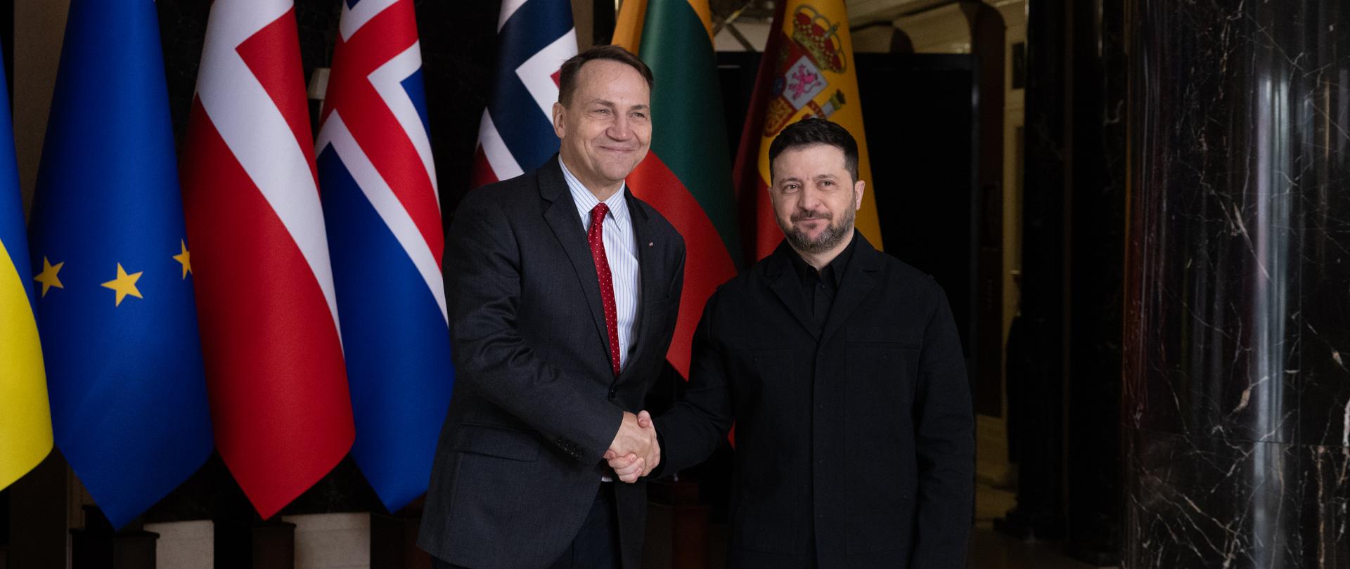 Na zdjęciu jest dwóch mężczyzn w ciemnych garniturach (Wicepremier, Minister Spraw Zagranicznych RP Radosław Sikorski oraz Prezydent Ukrainy Wołodymyr Zełenski). Podają sobie dłonie. Za nimi są flagi państw. 