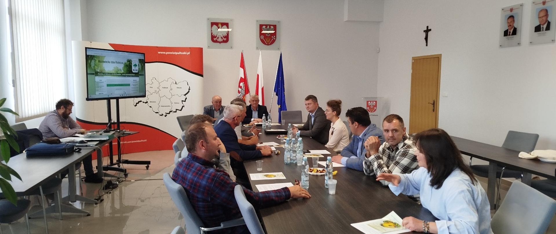 Sala konferencyjna podczas spotkania kilkunastu osób siedzących przy dużym stole. Na ścianie widoczne są flagi Polski i Unii Europejskiej, godła oraz ekran prezentacyjny. Uczestnicy prowadzą dyskusję.