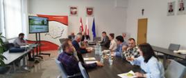 Sala konferencyjna podczas spotkania kilkunastu osób siedzących przy dużym stole. Na ścianie widoczne są flagi Polski i Unii Europejskiej, godła oraz ekran prezentacyjny. Uczestnicy prowadzą dyskusję.