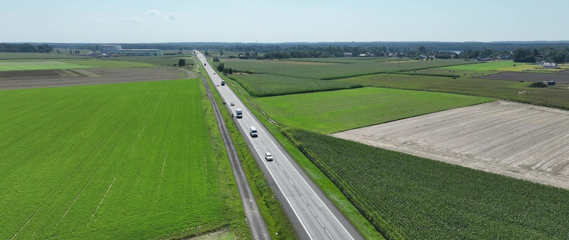 Droga krajowa nr 11 biegnąca między polami uprawnymi. w Tle po lewej zabudowania magazynowe i produkcyjne. Po prawej niewielka miejscowość z rzadkimi zabudowaniami.