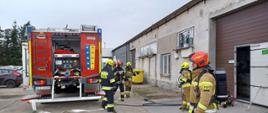 Na pierwszym planie widać strażaków w sprzęcie ochrony układu oddechowego po ewakuacji poszkodowanego z budynku objętego pożarem oraz kierującego działaniem ratowniczym. W tle stoi samochód pożarniczy na terenie przedsiębiorstwa.