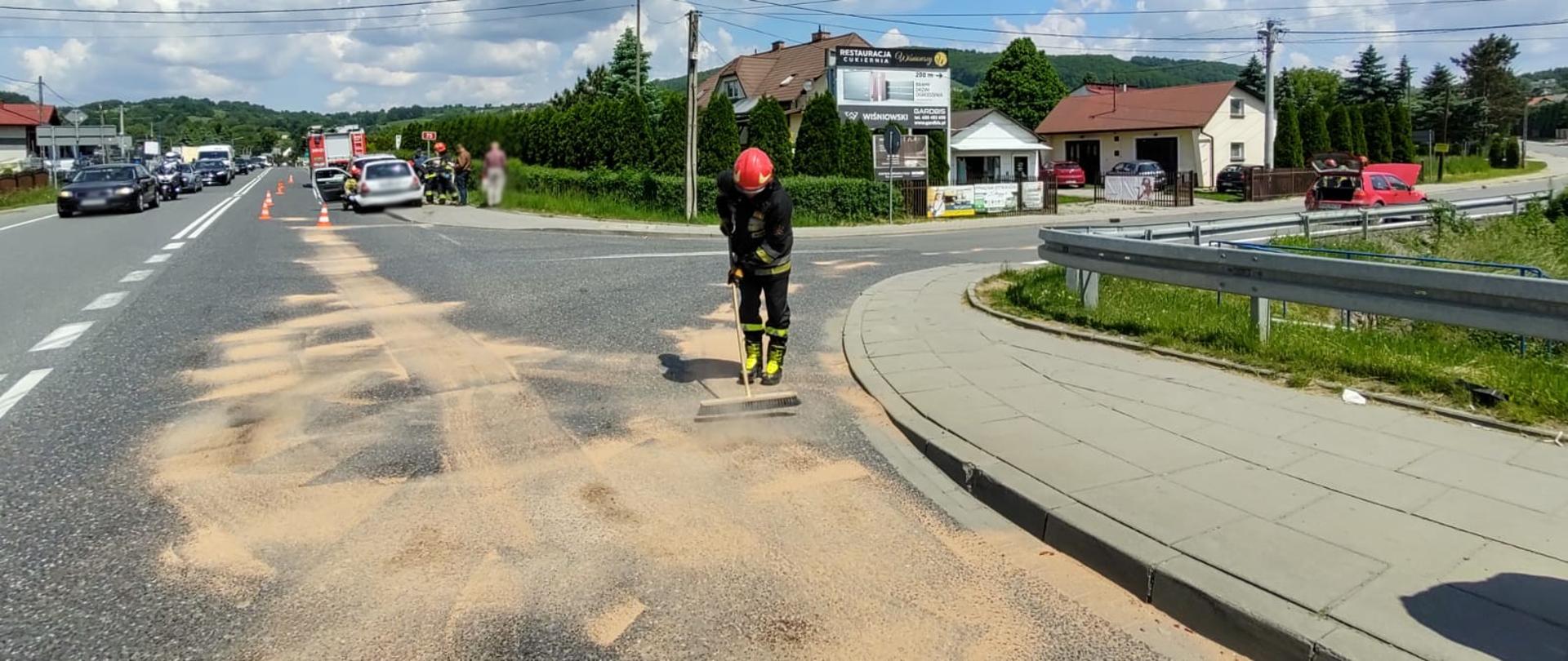 Wycieki zasypane sorbentem na drodze. Strażak miotłą rozprowadza sorbent. W tle widać stojące samochody.