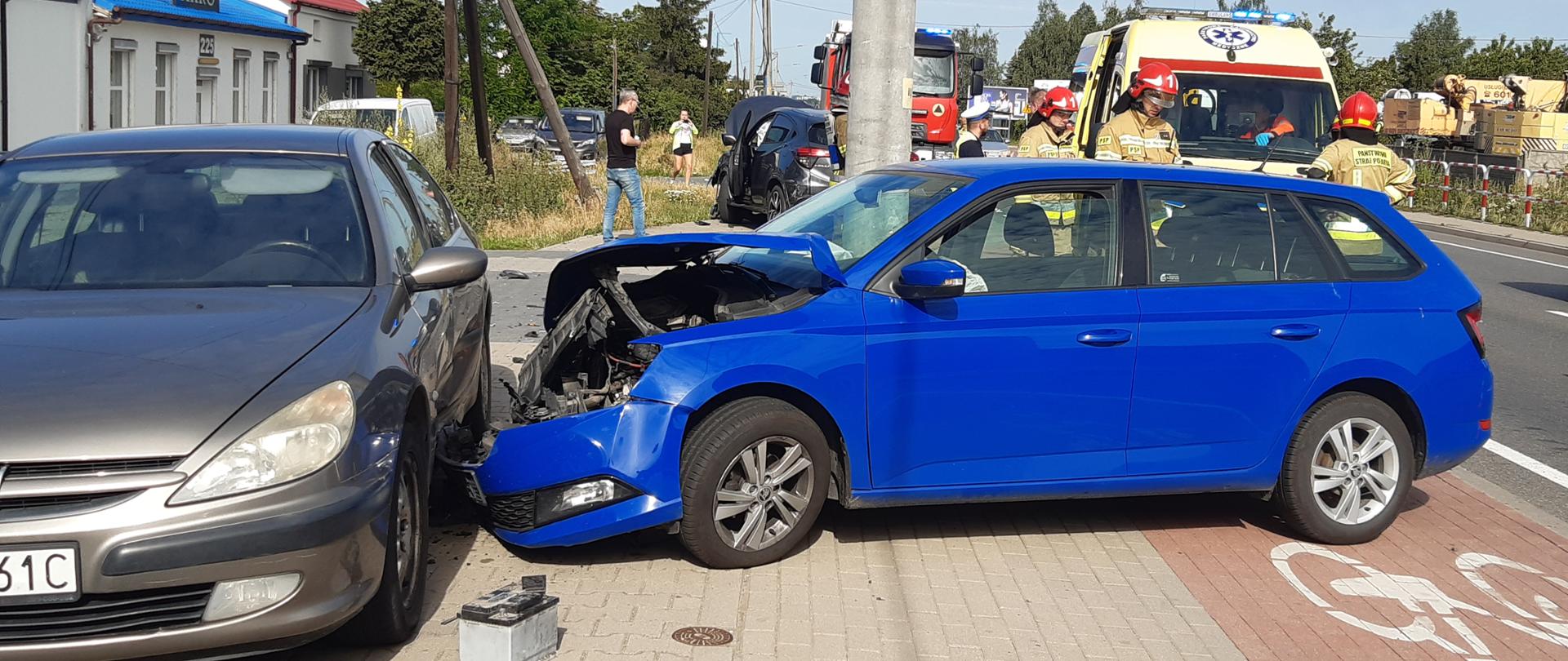 Na zdjęciu widać uszkodzone pojazdy. Są to samochody osobowe. Zdjęcie wykonane w ciągu dnia. Występuje zachmurzenie. Na drugim planie pojazdy służb mundurowych. Widać funkcjonariuszy.