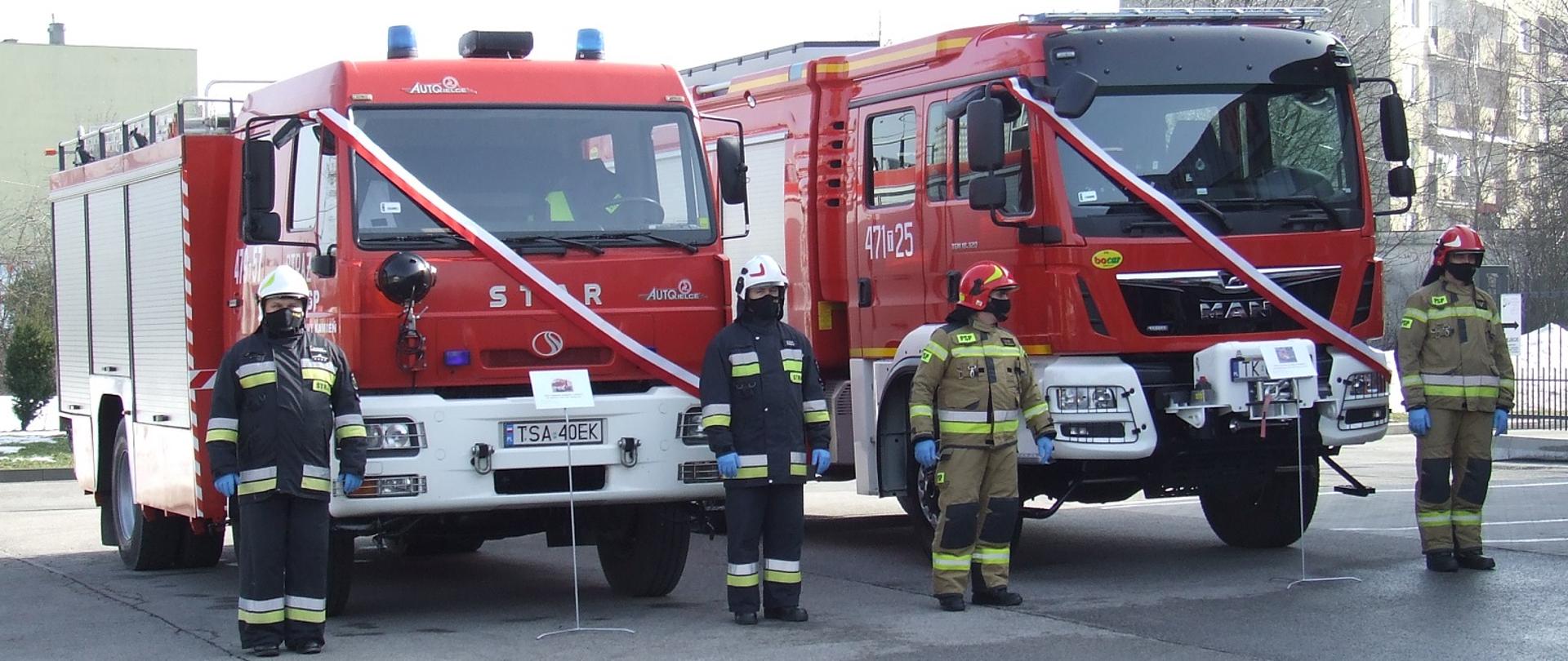 Zdjęcie zrobione na placu Komendy. Przedstawia dwa samochody strażackie. Na samochodach są zawieszone biało-czerwone szarfy. Przed każdym samochodem stoi dwóch strażaków, po prawej i po lewej stronie. Ubrani są w ubrania bojowe i hełmy a na twarzach mają ochronne maseczki.