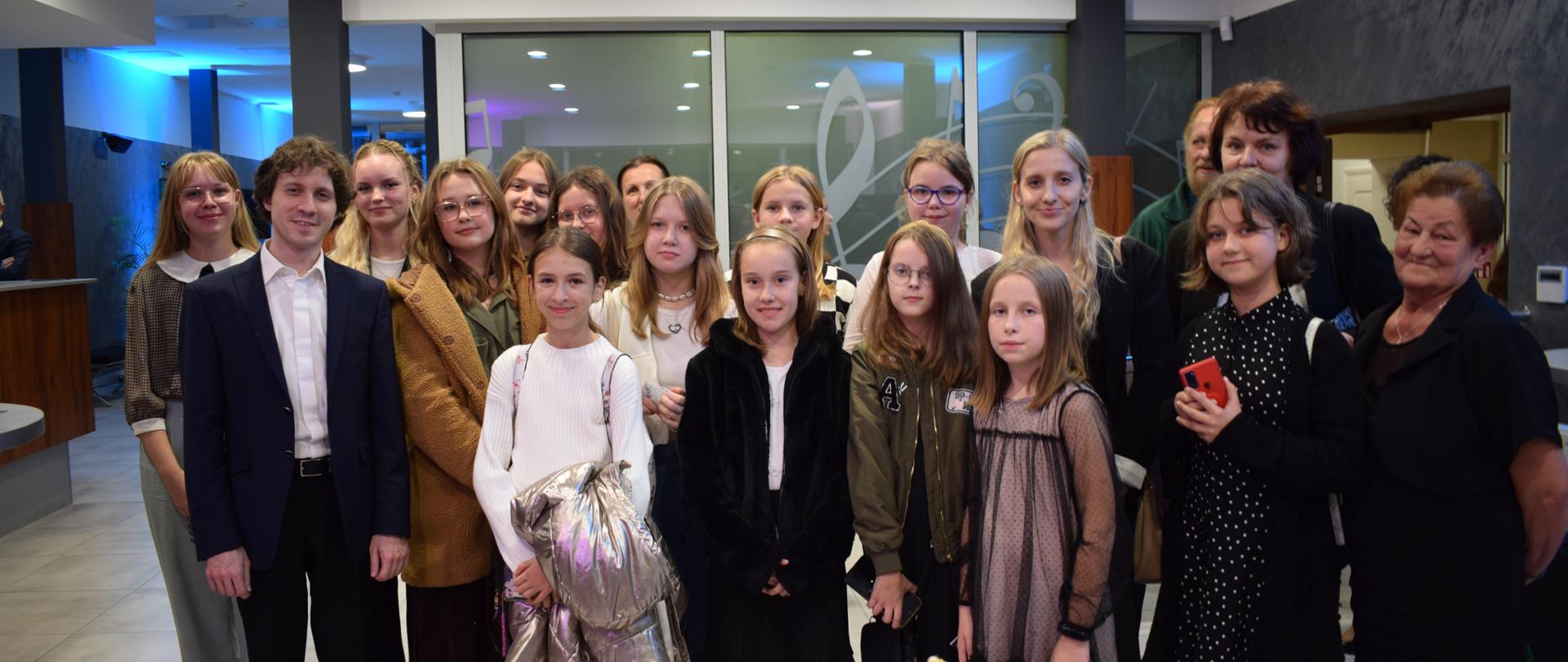 Zdjęcie przedstawia grupę uczniów i opiekunów po koncercie z pianistą Rafałem Blechaczem. Od lewej strony w pierwszym rzędzie stoi Maestro Rafał Blechacz ubrany w czarny garnitur i białą koszulę. W tle widać szklaną ścianę, składającą się z trzech zespolonych ze sobą części oddzielonych białymi szprosami. W każdą ramkę wstawiona jest mleczna szyba. N szkle przyklejona jest naklejka ze znakami muzycznymi (klucz muzyczny, nuta, klucz basowy). Przed ścianą widoczne są dwa filary w grafitowym kolorze. Z lewej strony widać korytarz prowadzący do sali koncertowej i światło odbijające się od ściany i sufitu z kolorze niebieskim. Z prawej natomiast przejście do drugiej części budynku. Ściany w tym pomieszczeniu od dołu częściowo pokryte są tynkiem strukturalnym w kolorze grafitowym. Reszta wraz z sufitem pomalowane są na biało. 