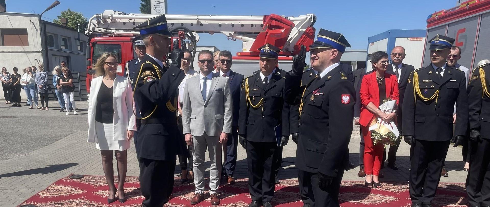 Zdjęcie przedstawia teren Jednostki Ratowniczo-Gaśniczej podczas obchodów Dnia Strażaka w Radziejowie. Na zdjęciu dowódca uroczystości ubrany w mundur galowy składa meldunek Kujawsko-Pomorskiemu Komendantowi Wojewódzkiemu Państwowej Straży Pożarnej o zakończeniu uroczystego apelu. Na drugim planie przedstawiciele samorządu terytorialnego oraz zaproszeni goście oraz wozy straży pożarnej.