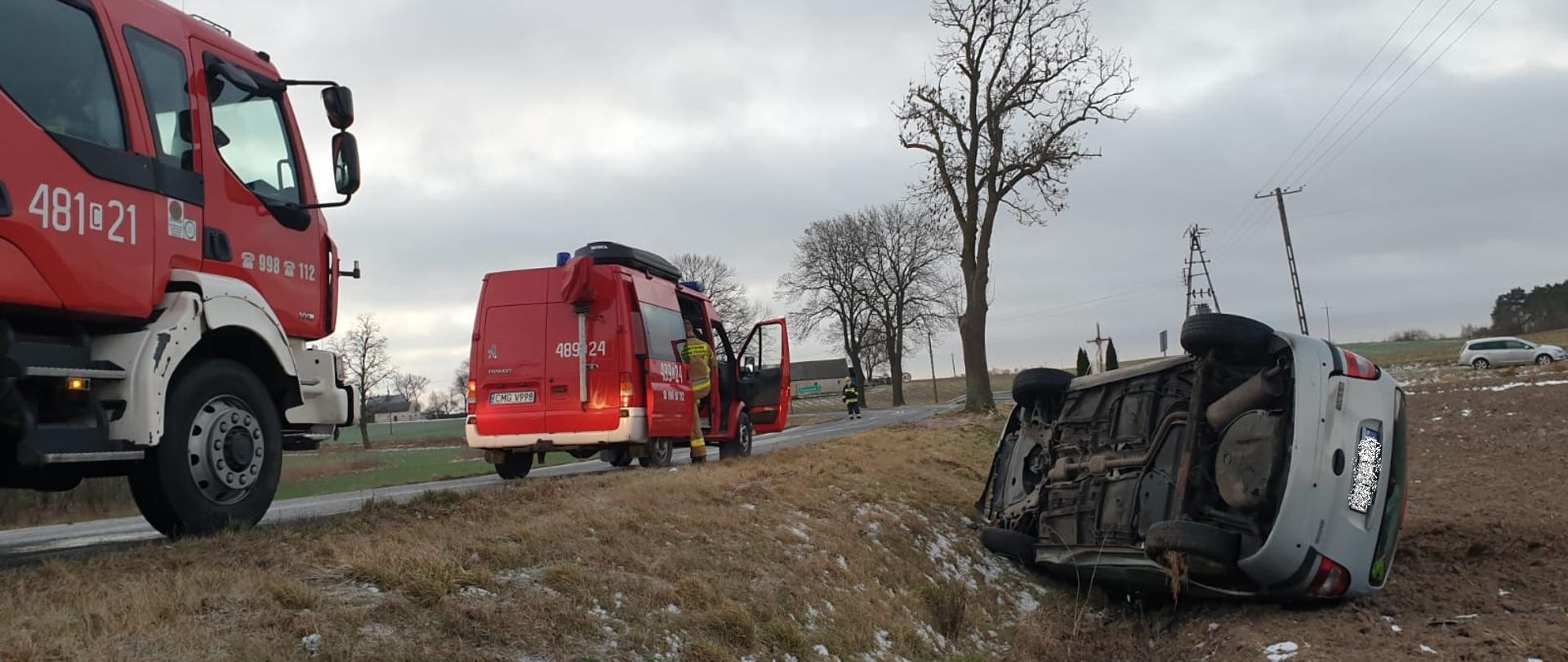 Samochód osobowy koloru srebrnego leży w polu na prawym boku. Z lewej strony 2 pojazdy pożarnicze