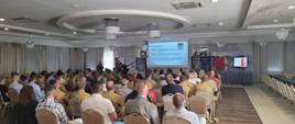 Zdjęcie przedstawia strażaków oraz osoby cywilne podczas seminarium. Osoby siedzą na krzesełkach na sali , natomiast prelegent omawia temat.