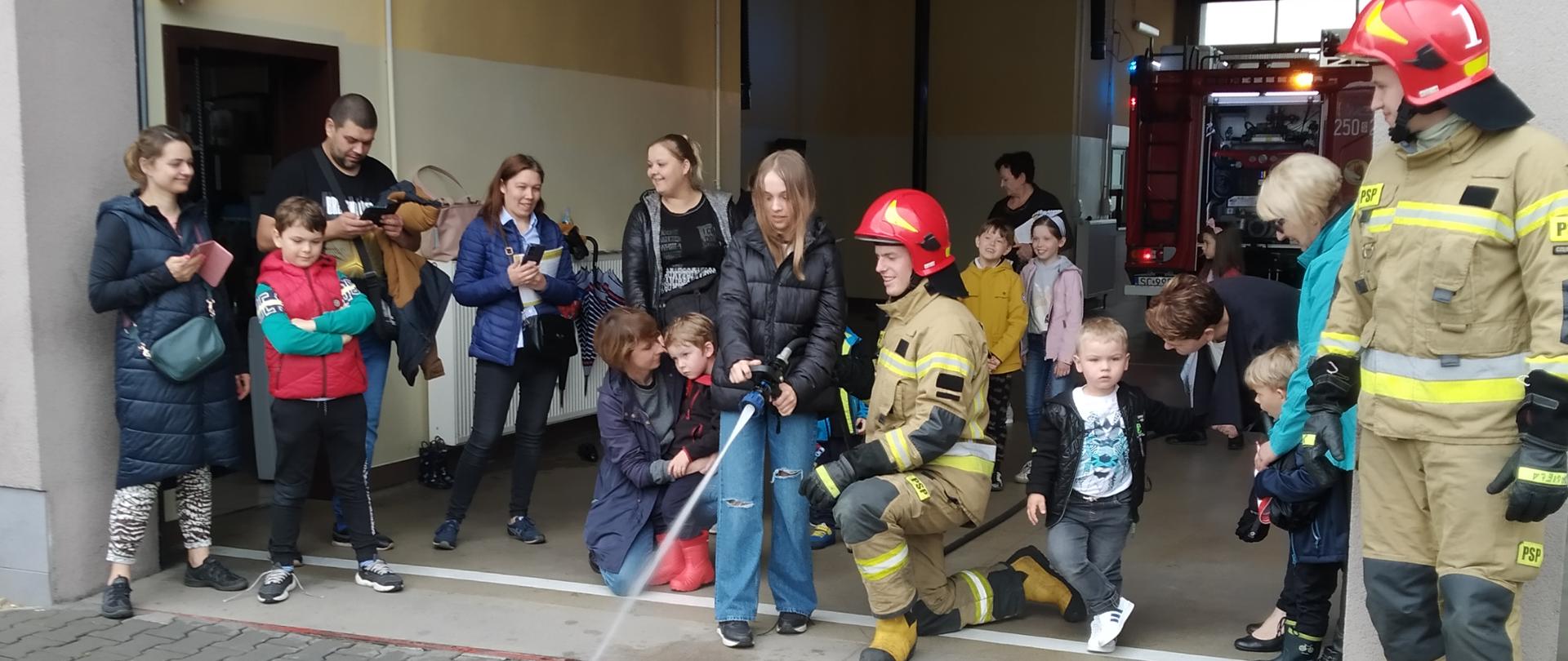 Na zdjęciu wnętrze garażu Jednostki Ratowniczo-Gaśniczej Centralnej Szkoły PSP, w którym znajduje się grupa dzieci wraz z opiekunami. Na środku dziewczynka, w asyście strażaka, trzymając prądownicę kieruje strumieniem wody