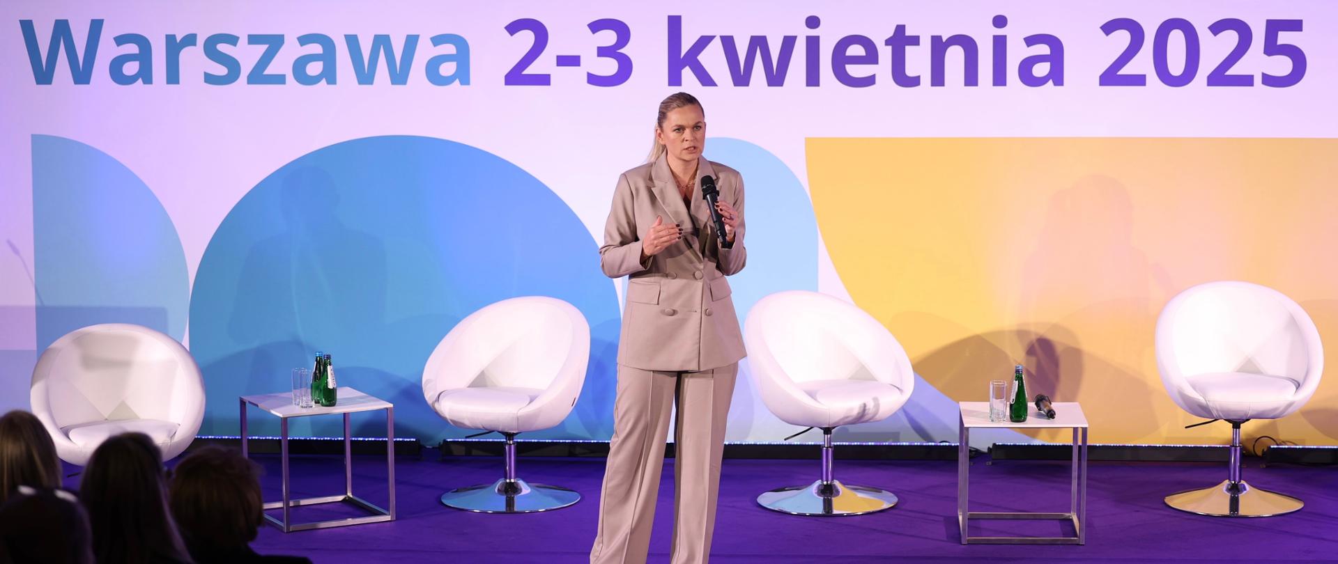 Na tle kolorowej ścianki z napisem New Education Forum Warszawa 2-3 kwietnia 2025 stoi Barbara Nowacka i mówi do mikrofonu, który trzyma w ręku. Za nią cztery fotele i dwa stoliki.