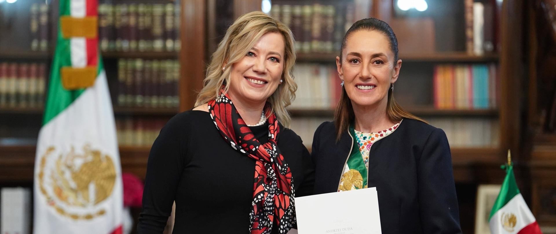 En la foto aparecen dos mujeres sonrientes juntas en una sala elegante y representativa. La persona de la izquierda es la embajadora de Polonia en México, Agnieszka Frydrychowicz Tekieli, y la de la derecha es la presidenta de México, Claudia Sheinbaum. Ambas visten de gala y sostienen en sus manos un maletín blanco con un documento: las cartas credenciales. Al fondo se ven estanterías con libros y banderas de México. La escena tiene un carácter solemne y oficial.