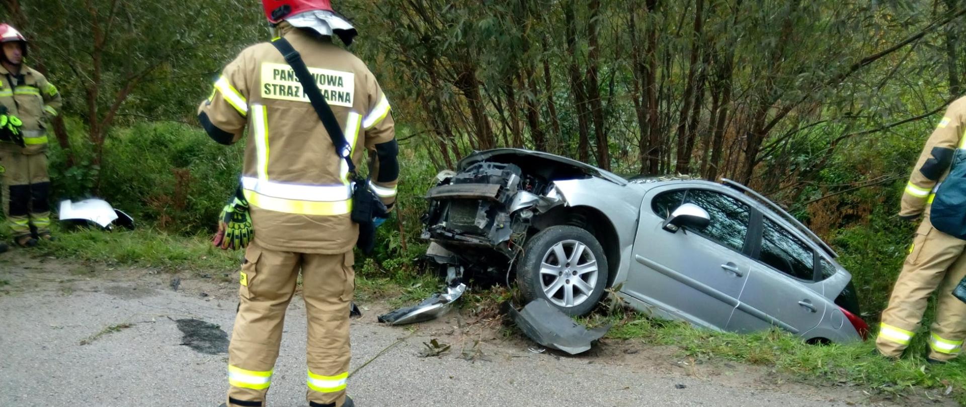 Zdjęcie przedstawia samochód osobowy, który wypadł z drogi i znajduje się w przydrożnym rowie. Wokół pojazdu stoi trzech ratowników. 