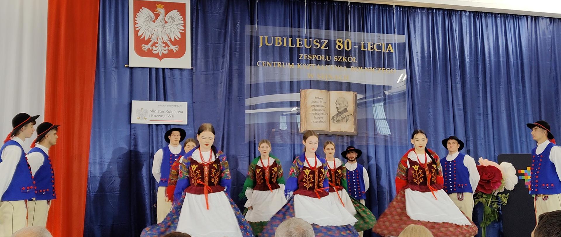 Na scenie tańczy zespół ludowy, w tle dekoracja z napisem "Jubileusz 80-lecia Zespołu Szkół Centrum Kształcenia Rolniczego w Sejnach".