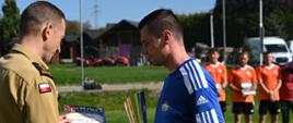 Uroczysta dekoracja uczestników zawodów na boisku sportowym. Z lewej strony strażak w piaskowym mundurze służbowym wręcza dyplom za zajęcie II miejsca oraz duży, srebrno-złoty puchar zawodnikowi w niebieskiej koszulce Adidas. W tle na trawie stoją pozostali zawodnicy w pomarańczowych koszulkach.