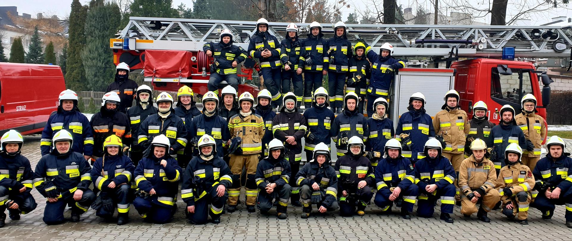 Kilkadziesiąt osób w ubraniach bojowych straży pożarnej, w białych hełmach stojących w trzech rzędach. W tle czerwony samochód pożarniczy z drabiną mechaniczną. 