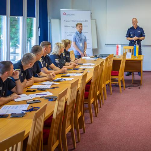 W sali lekcyjnej grupa mężczyzn i kobiet siedzi przy ustawionych naprzeciwko siebie dwóch rzędach stołów i patrzy się na prowadzących szkolenie trzech mężczyzn stojących przed pustym ekranem, tablicą flipchart i banerem z logo Polskiej pomocy