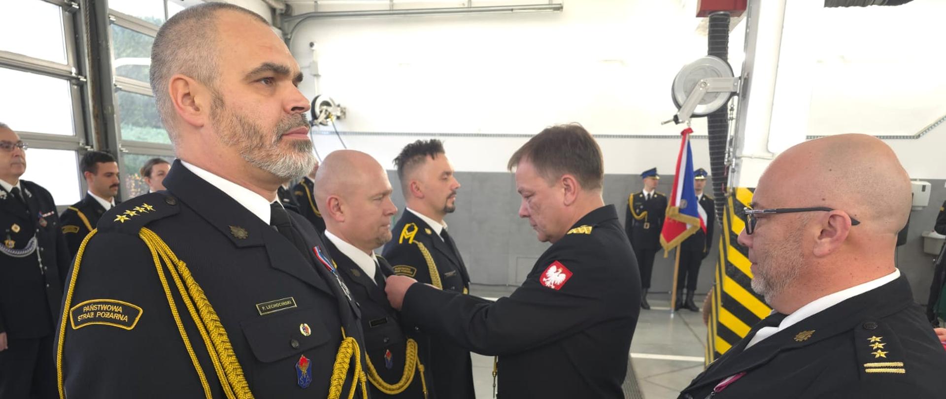 Uroczysta zbiórka z okazji Święta Niepodległości - przed frontem pododdziału wystąpili trzej funkcjonariusze, Komendant Wojewódzki dekoruje strażaka w środku odznaczeniem. Strażacy w umundurowaniu galowym. Po prawej Komendant Powiatowy asystujący przy odznaczaniu strażaków. W tle poczet sztandarowy komendy Powiatowej PSP w Krapkowicach. 