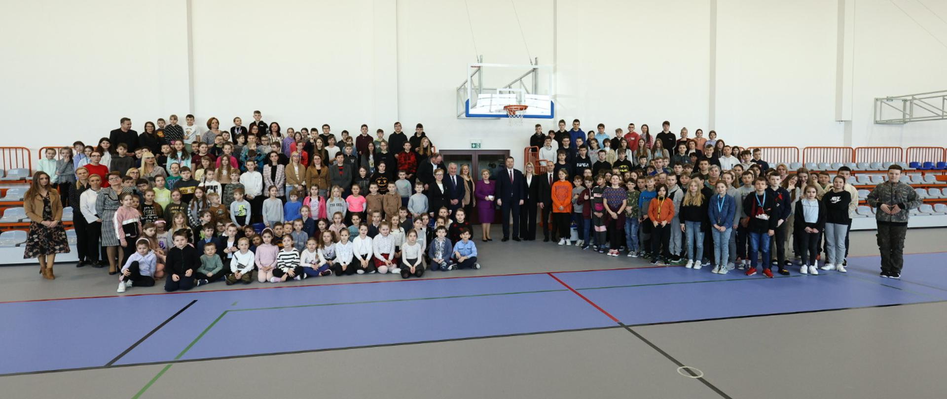 Widok na halę sportową, na szarej podłodze stoją dwie duże grupy kolorowo ubranych dzieci.