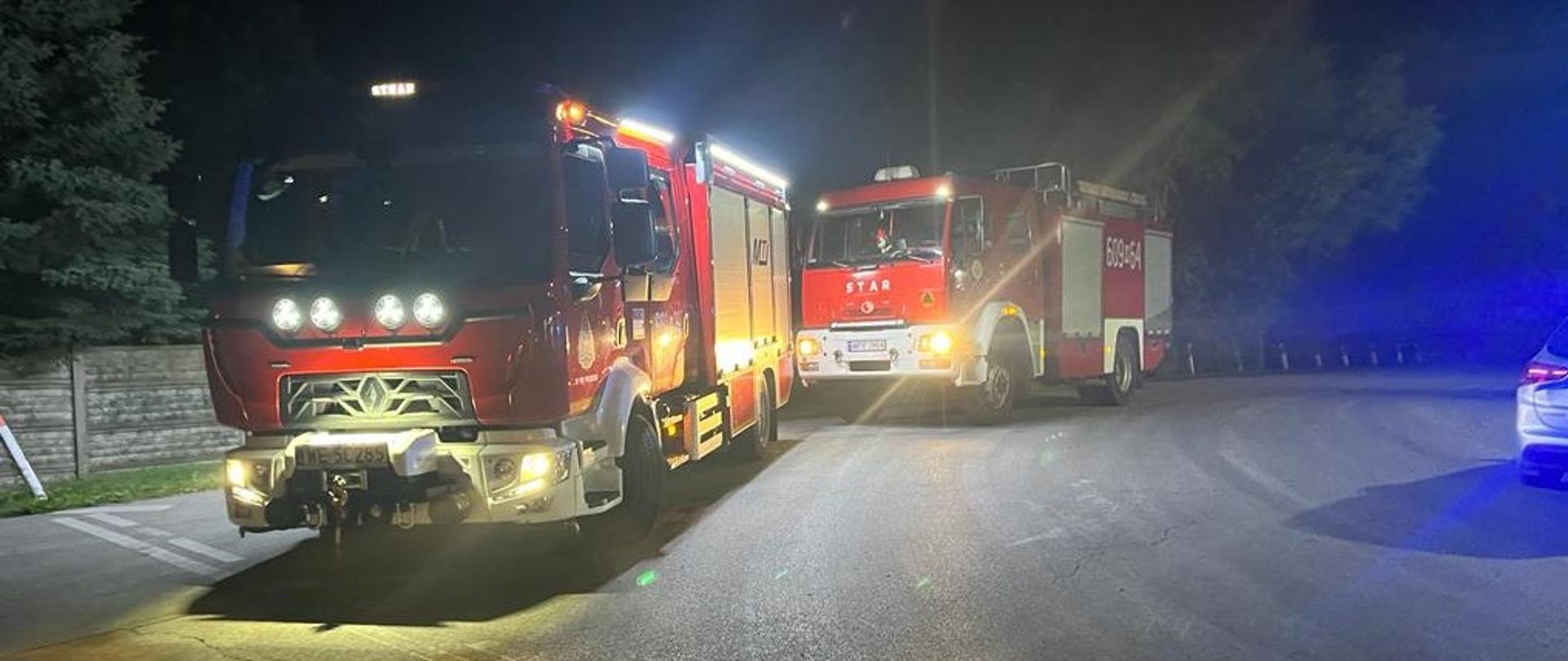 
Zdjęcie przedstawia dwa czerwone wozy strażackie stojące na drodze w nocy. Na pierwszym planie, po lewej, znajduje się większy wóz z włączonymi światłami, w tym światłami awaryjnymi, oraz widoczną rejestracją "WE 5C285". Za nim, po prawej stronie, stoi drugi, nieco mniejszy wóz strażacki z numerem "609-" i napisem "STAR". Obok drugiego wozu widoczny jest fragment trzeciego pojazdu z niebieskimi światłami, prawdopodobnie policyjnego lub ratunkowego. Otoczenie jest ciemne, oświetlone jedynie przez światła pojazdów, co sugeruje nocną interwencję.