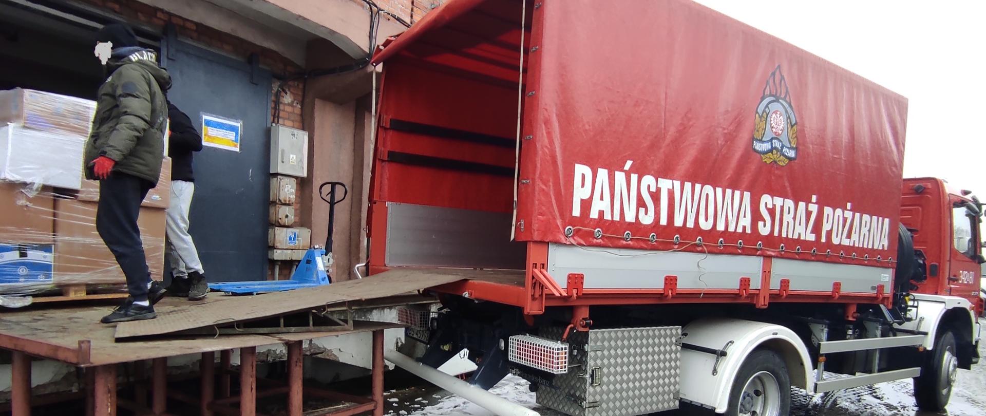 Na zdjęciu widać samochód straży pożarnej na który ładowane są palety z paczkami oraz dwie osoby
