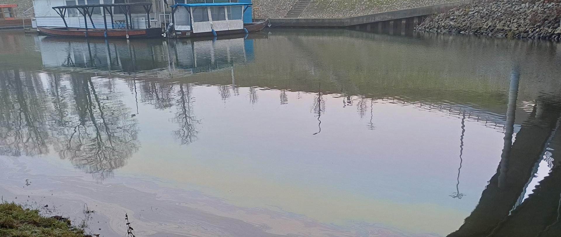 Spokojna powierzchnia wody z widocznym filmem substancji ropopochodnych. W tle zacumowane barki i umocniony brzeg kanału.