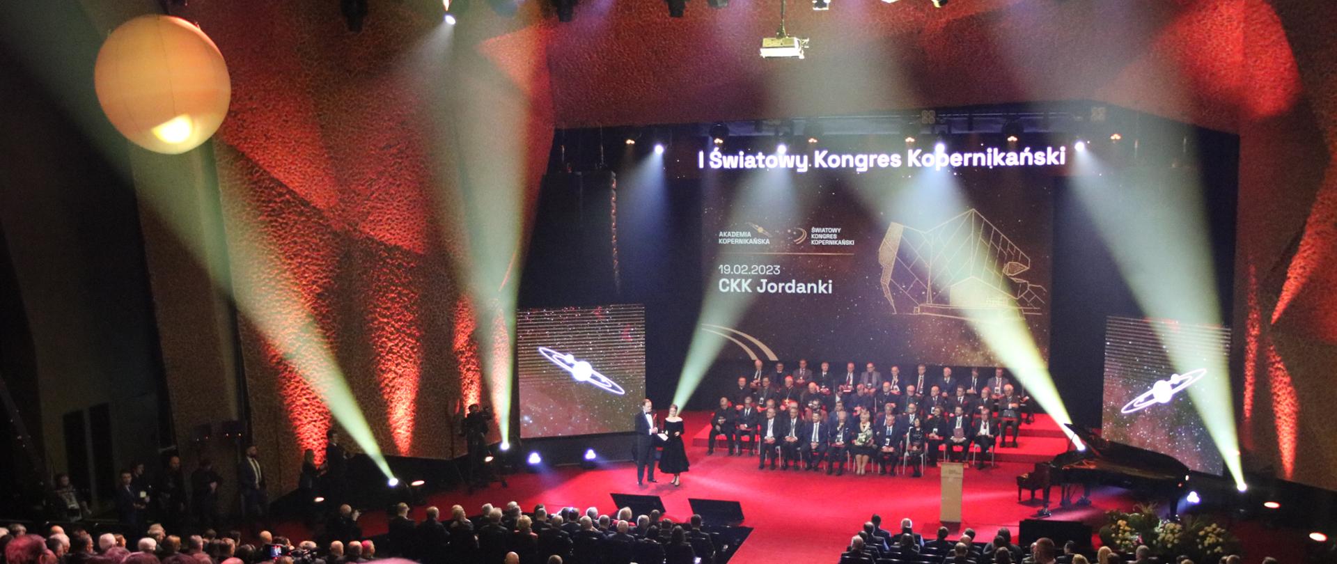 Widok z tyłu na wypełnioną ludźmi salę, z przodu na podwyższeniu siedzi na krzesłach duża grupa ludzi, za nimi wielki ekran z napisem I Światowy Kongres Kopernikański.