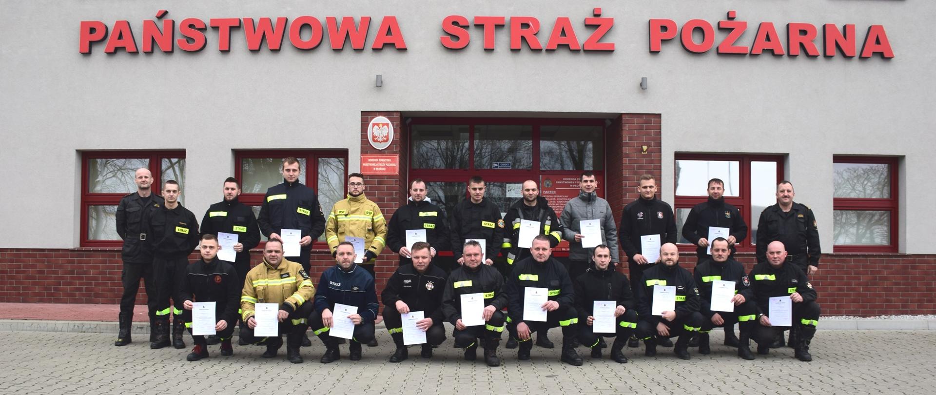 Grupa strażaków w ubraniach koszarowych pozuje przed budynkiem z napisem: Państwowa Straż Pożarna.