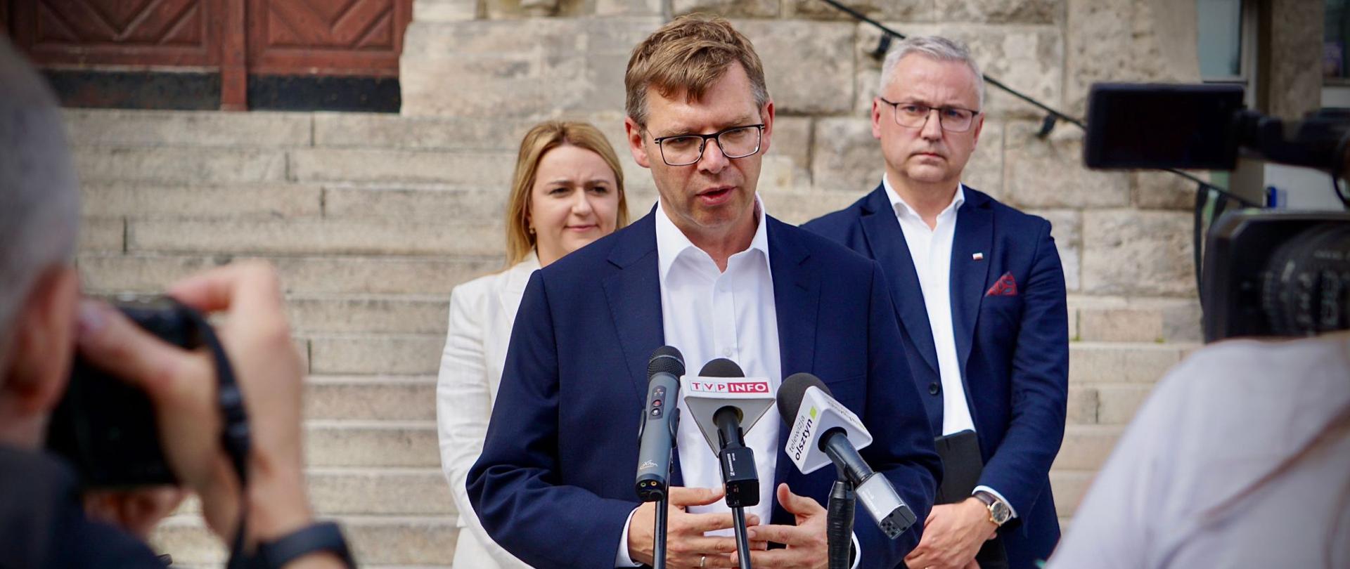 Konferencja Pasowa: Wojewoda Warmińsko-Mazurski Artur Chojecki stoi przed mikrofonem i przemawia a za nim stoi kobieta i mężczyzna 