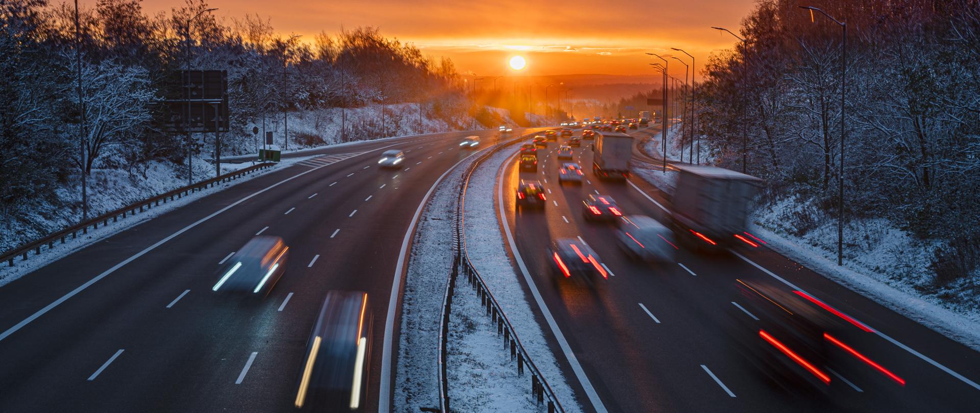 Zimowy poranek na wielopasmowej autostradzie. Na drodze w obu kierunkach jedzie wiele samochodów, widoczne są smugi świateł przez długi czas naświetlania. Po bokach trasy znajdują się ośnieżone drzewa, a na horyzoncie widać wschodzące słońce, które oświetla niebo ciepłymi barwami.