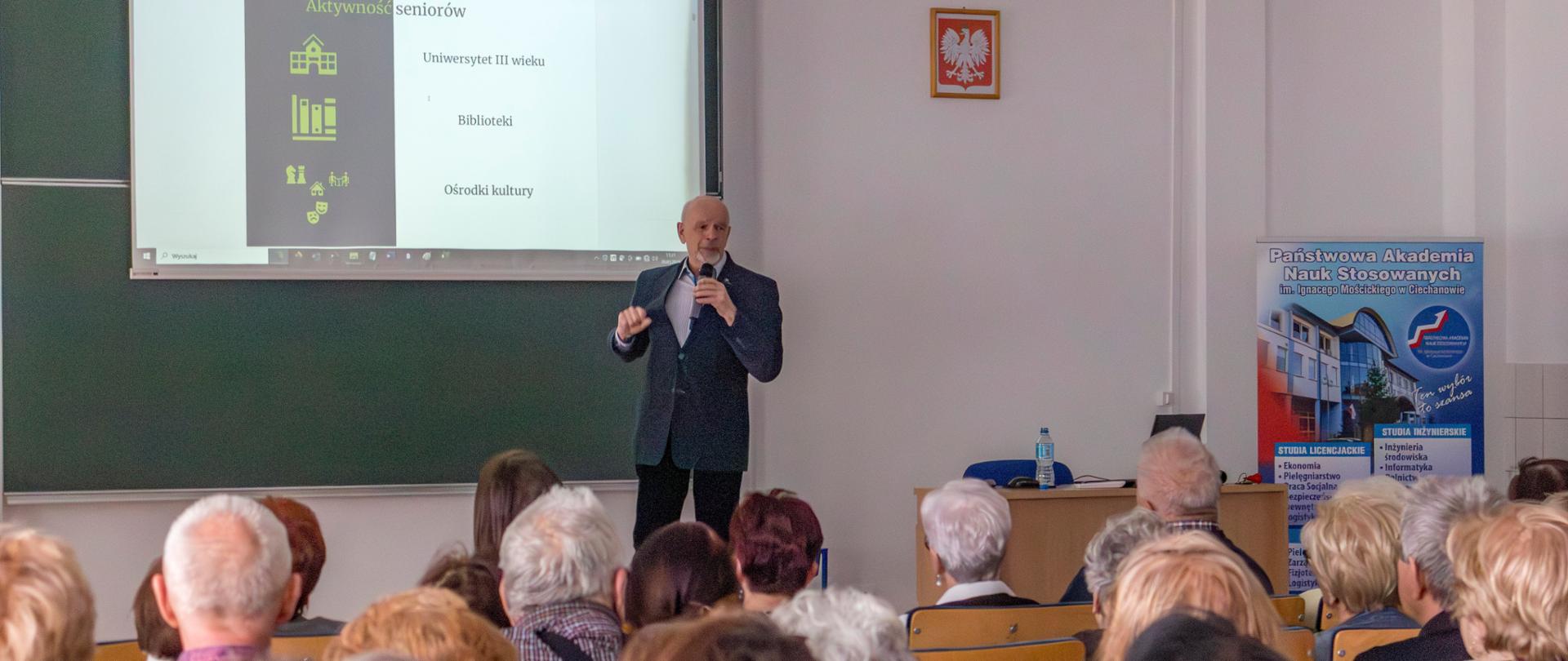 Grupa osób siedząca na sali wykładowej, słuchają wykładowcy mówiącego do mikrofonu na tle wyświetlanej prezentacji