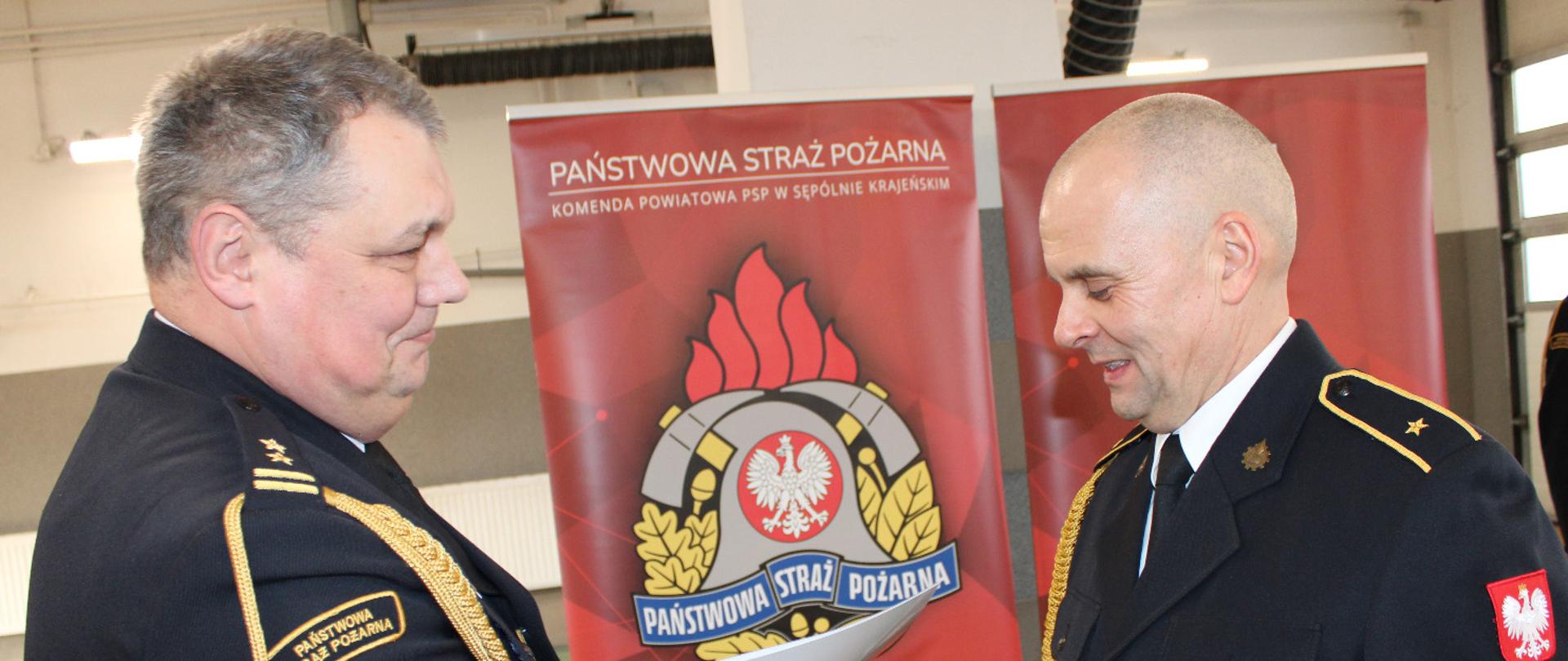 Komendant brygadier Marcin Mazurkiewicz w mundurze wyjściowym ściska dłoń i wręcza dokument odchodzącemu na zaopatrzenie emerytalne