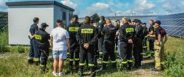  Strażacy podczas zapoznania z infrastrukturą farmy fotowoltaicznej. Wszyscy stoją na trawie przodem do budynku stacji transformatorowej, szkolenie prowadzi wykonawca instalacji. W tle panele fotowoltaiczne oraz błękit nieba z białymi chmurami.