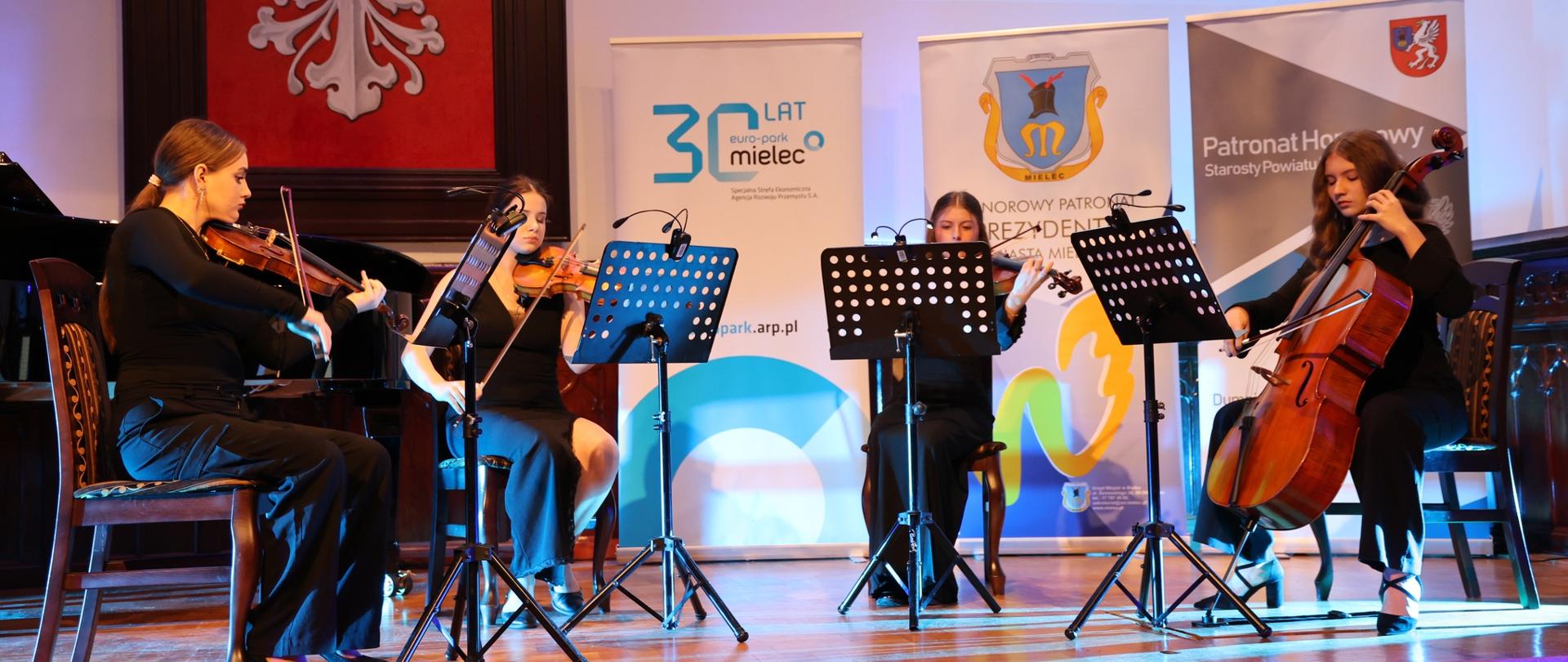 Cztery młode instrumentalistki grające w kwartet smyczkowy na scenie Sali Królewskiej; trzy skrzypaczki i wiolonczelistka występują przed publicznością na tle banerów wydarzenia i dużego godła z orłem.