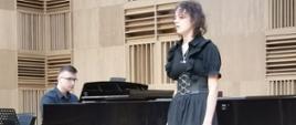 Na scenie stoi młoda dziewczyna o jasnych, krótkich włosach. Śpiewa. Przy fortepianie akomapniuje jej mężczyzna.